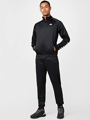 Man wearing a black tracksuit with logo, hands in pockets.