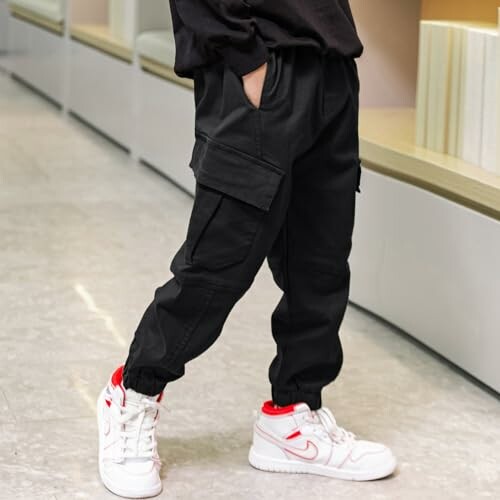 Child in black cargo pants and white sneakers with red accents standing indoors.