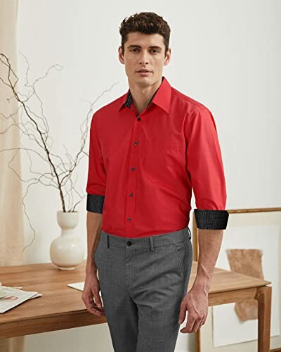 Man in red shirt and gray pants standing by a wooden table.